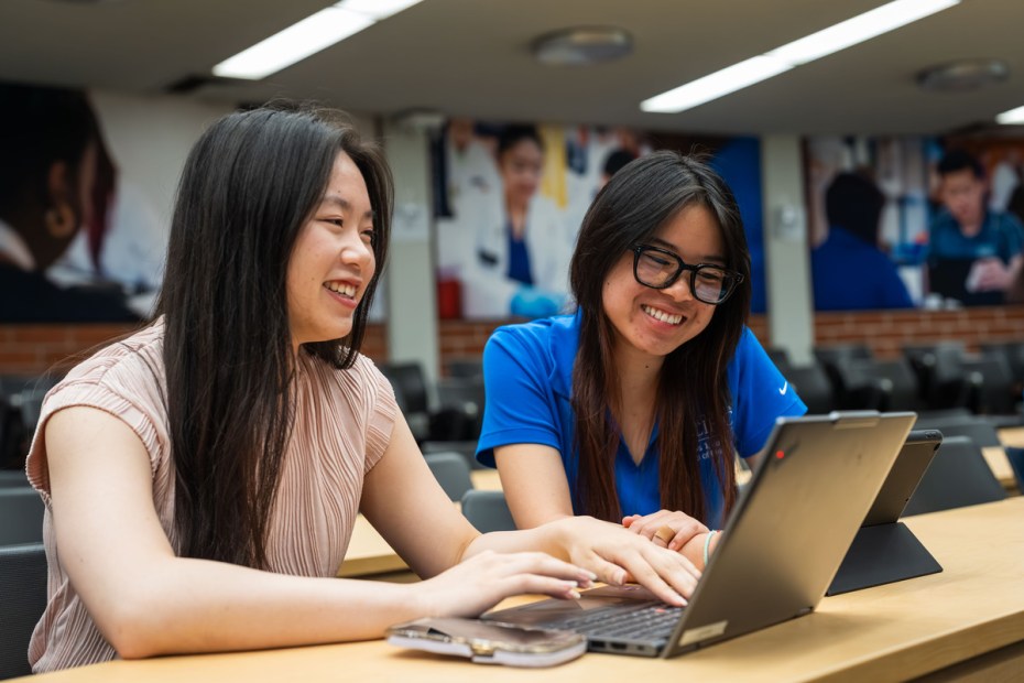 Two students using their laptops in a classroom, both smiling.